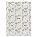 A rectangular fabric item, likely a baby blanket or towel, featuring a repeating pattern of green cartoon dinosaurs, pink and beige flower clusters, and the name 'REMY' printed in brown on a white background.