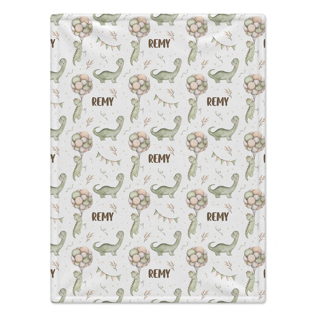 A rectangular fabric item, likely a baby blanket or towel, featuring a repeating pattern of green cartoon dinosaurs, pink and beige flower clusters, and the name 'REMY' printed in brown on a white background.