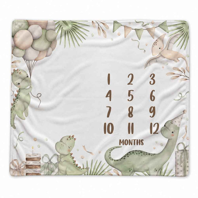 A square baby blanket with a white background featuring watercolor-style dinosaur illustrations, balloons, palm leaves, bunting, a birthday cake, and a printed monthly countdown from 1 to 12 labeled 'MONTHS'.