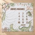 A personalized dinosaur-themed baby milestone blanket laid flat on a light wood floor. The white blanket features illustrations of green dinosaurs, balloons, a birthday cake, and a pterodactyl, with the text 'JAMES MICHAEL' at the top and a '12 MONTHS' timeline on the right.