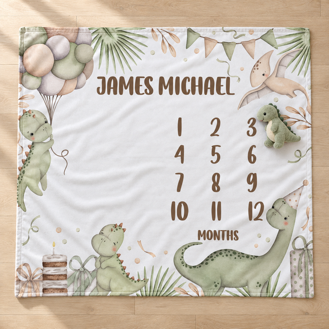 A personalized dinosaur-themed baby milestone blanket laid flat on a light wood floor. The white blanket features illustrations of green dinosaurs, balloons, a birthday cake, and a pterodactyl, with the text 'JAMES MICHAEL' at the top and a '12 MONTHS' timeline on the right.