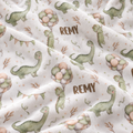 A close-up of a soft white fabric with a repeating dinosaur print in muted green and peach, plus the name 'REMY' in brown lettering.