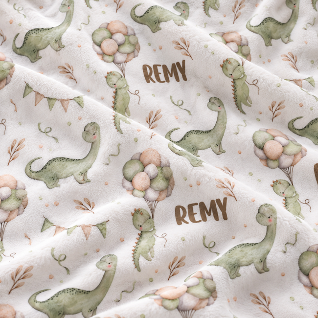 A close-up of a soft white fabric with a repeating dinosaur print in muted green and peach, plus the name 'REMY' in brown lettering.