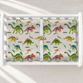 A white changing table with a personalized changing pad cover featuring a colorful pattern of cartoon dinosaurs on a warm cream background with the word 'CAIDEN' printed below each dinosaur.