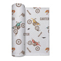 A rolled baby swaddle blanket with a western and dirt-bike pattern, shown next to the flat sheet. The design features brown and beige western boots, checkered flags, wheat stalks, and motocross bikes in red, yellow, and green on a light gray background with the name 'CARTER' printed in brown.