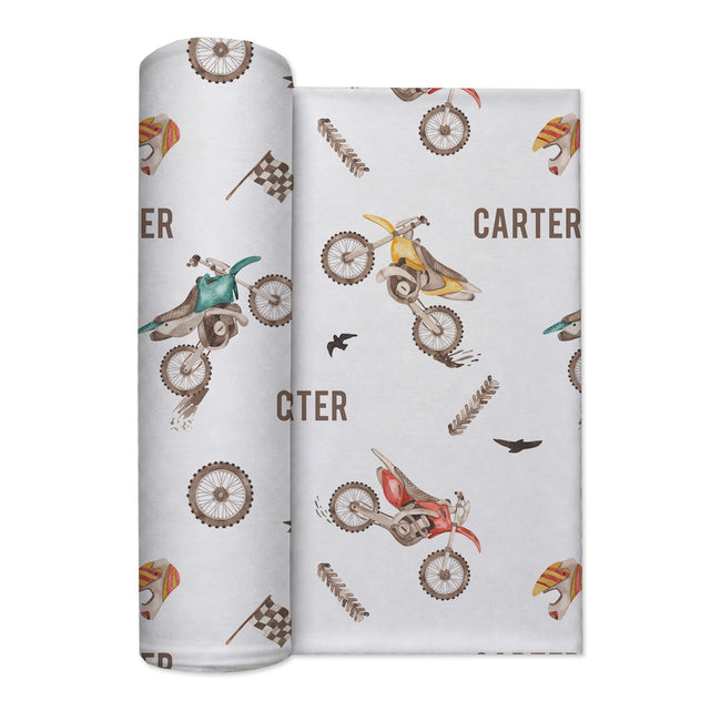 A rolled baby swaddle blanket with a western and dirt-bike pattern, shown next to the flat sheet. The design features brown and beige western boots, checkered flags, wheat stalks, and motocross bikes in red, yellow, and green on a light gray background with the name 'CARTER' printed in brown.
