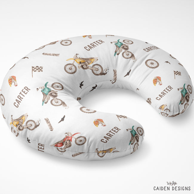 A U-shaped nursing pillow with a white background and a repeating pattern of dirt bikes, checkered flags, birds, and the name 'CARTER' in brown text. The brand 'CAIDEN DESIGNS' is visible in the bottom right corner.