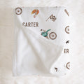 A white personalized baby blanket with a vintage bicycle theme, featuring the name 'CARTER' and illustrations of bicycles, birds, and a checkered flag, laid flat on a white faux fur background.