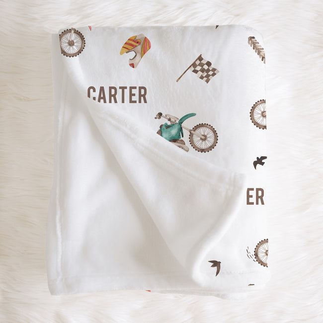 A white personalized baby blanket with a vintage bicycle theme, featuring the name 'CARTER' and illustrations of bicycles, birds, and a checkered flag, laid flat on a white faux fur background.