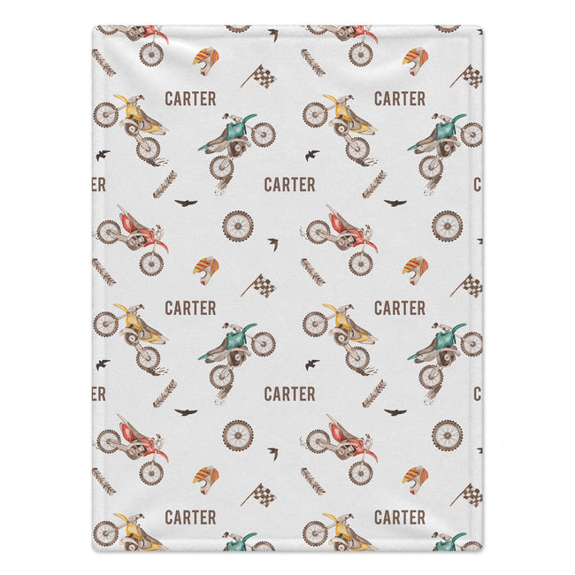A rectangular white blanket featuring a repeating pattern of vintage dirt bikes, checkered flags, birds, and the name 'CARTER' printed in brown.