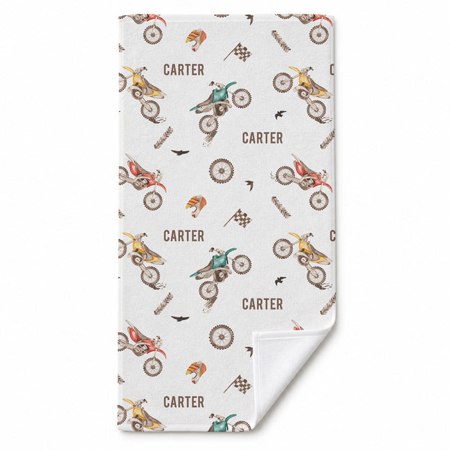 A rectangular towel with a white background and a repeating pattern of vintage dirt bikes, racing flags, checkered flags, and the name 'CARTER' in brown. The bottom right corner is folded over to show the white reverse side.