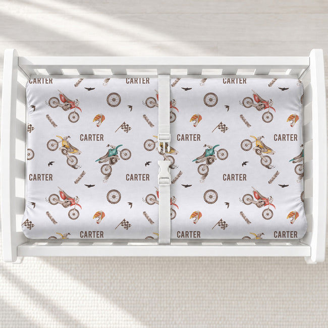A white changing table with a personalized changing pad cover featuring dirt bike and motorcycle pattern printed with the name 'CARTER' in brown.