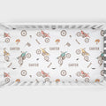 A white crib bumper sticker or bedding insert featuring a repeating pattern of vintage dirt bikes, checkered flags, birds, and the name 'CARTER' in brown, displayed inside a white wooden crib.