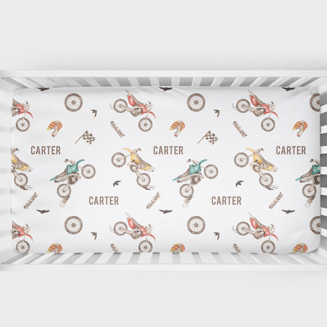 A white crib bumper sticker or bedding insert featuring a repeating pattern of vintage dirt bikes, checkered flags, birds, and the name 'CARTER' in brown, displayed inside a white wooden crib.