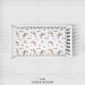 A white wooden crib shown from above on a light whitewashed wood background, with a printed sheet inside featuring a repeating pattern of dirt bikes, wheels, and the name Carter. The brand name 'CAIDEN DESIGNS' appears below the crib.