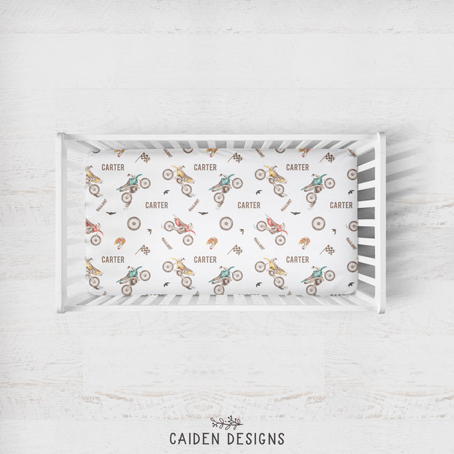 A white wooden crib shown from above on a light whitewashed wood background, with a printed sheet inside featuring a repeating pattern of dirt bikes, wheels, and the name Carter. The brand name 'CAIDEN DESIGNS' appears below the crib.
