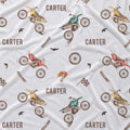 A close-up of a white fabric with a repeating printed pattern featuring dirt bikes, the name 'CARTER', birds, flags, and small brown and red accents.