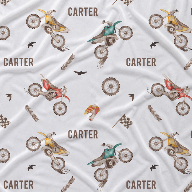 A close-up of a white fabric with a repeating printed pattern featuring dirt bikes, the name 'CARTER', birds, flags, and small brown and red accents.