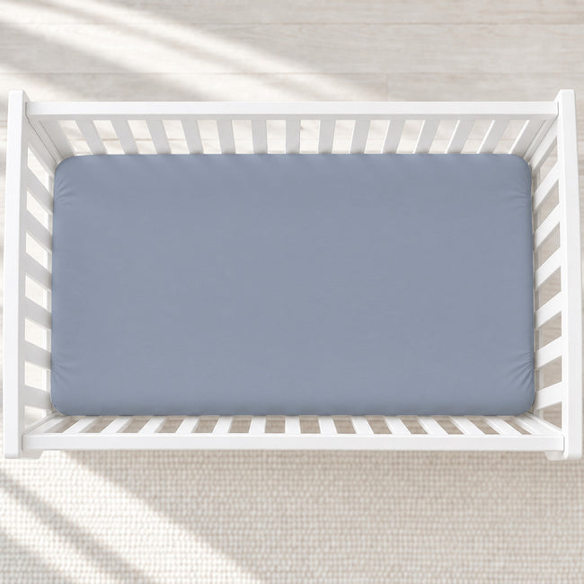 A top-down view of a light blue fitted crib mattress inside a white wooden crib, set on a textured cream rug with soft diagonal sunlight shadows.