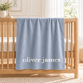 A light blue personalized baby blanket with the name 'oliver james' printed in white, draped over a wooden crib in a bright nursery setting.