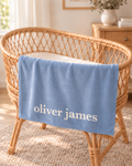A personalized light blue baby blanket with the name 'oliver james' in white script, draped over a natural wicker bassinet in a softly lit nursery setting.