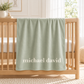 A pale sage green baby blanket with the personalized name 'michael david' printed in white at the bottom, draped over a light wood crib in a bright nursery.