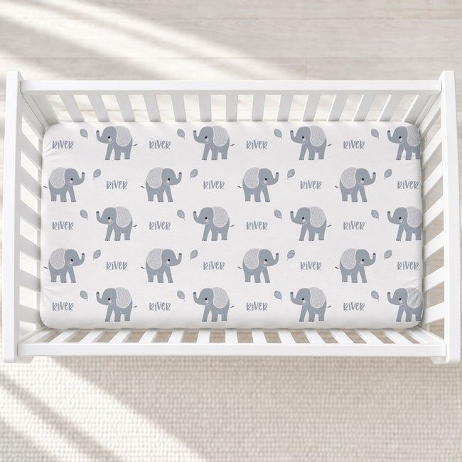 A white crib mattress fitted sheet with a repeating pattern of gray baby elephants and the word 'river' in small black text, shown inside a white wooden crib.