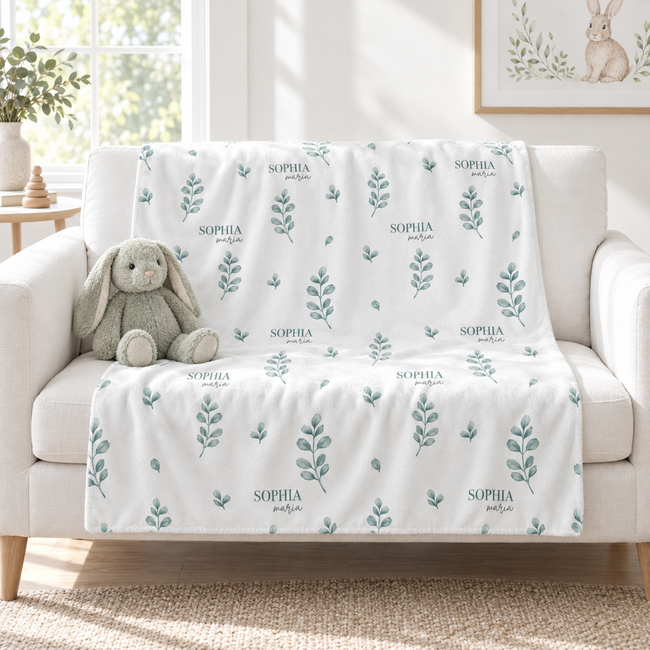 A white throw blanket with a repeating pattern of sage green leafy stems and the name 'SOPHIA printed in small dark text, draped over a light beige sofa in a bright living room. A plush bunny toy sits on the sofa.
