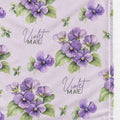 A close-up of a fabric or wallpaper pattern featuring watercolor-style purple violets with green leaves on a pale lavender background, with the text 'Violet MAE' printed in script and serif lettering.