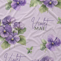 Pale lavender fabric with purple violet flowers, green leaves, and the name Violet Mae in purple script and green lettering.