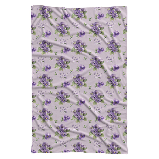 Pale lavender swaddle blanket with purple violet flowers, green leaves, and the name Violet Mae in purple and green lettering.