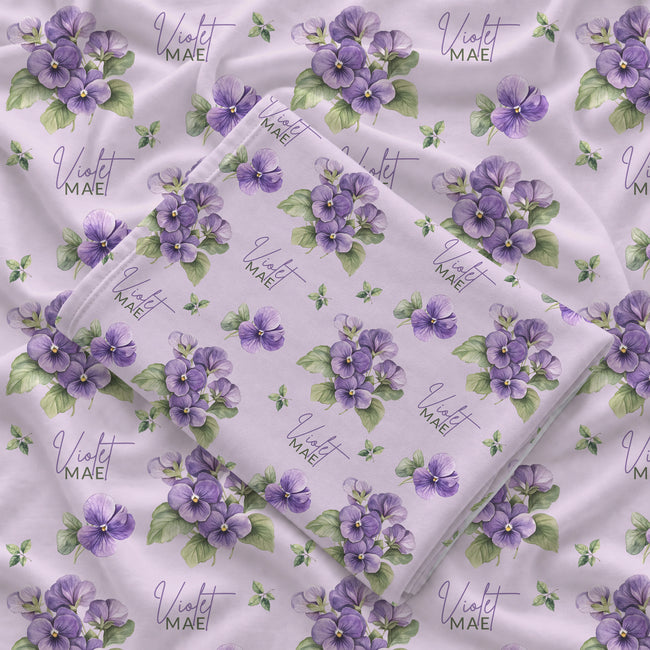 Pale lavender swaddle blanket with purple violet flowers, green leaves, and the name Violet Mae in purple and green lettering.