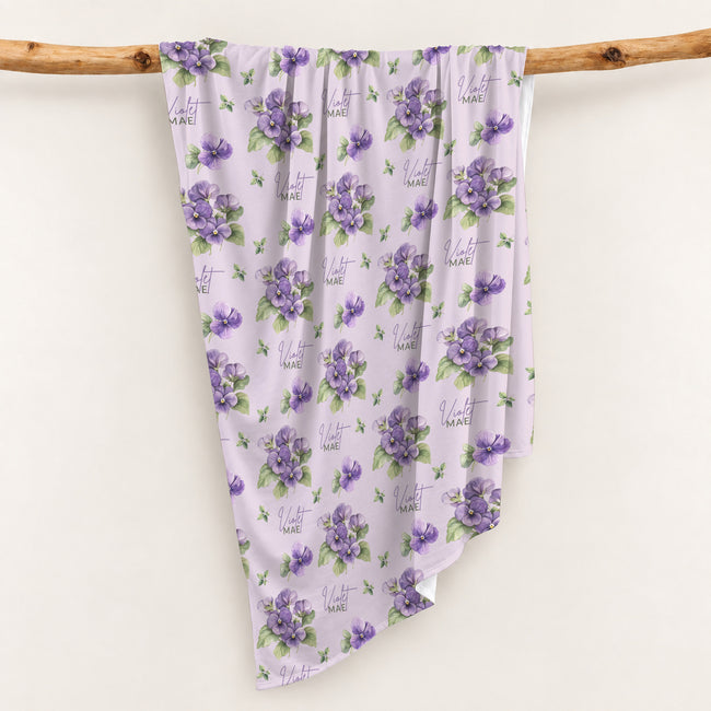 Pale lavender swaddle blanket with purple violet flowers, green leaves, and the name Violet Mae in purple and green lettering.