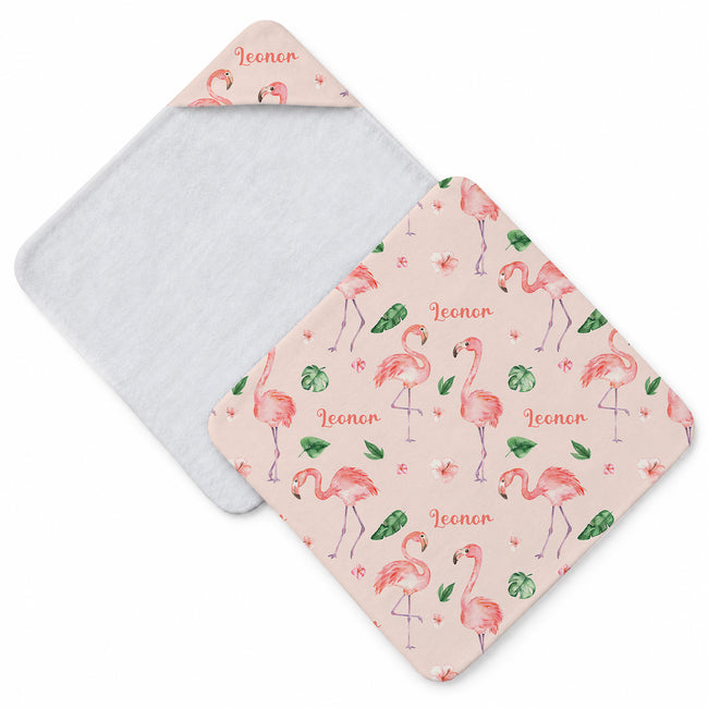 Pale blush hooded towel with pink flamingos, green tropical leaves, small flowers, and the name Leonor in coral script.