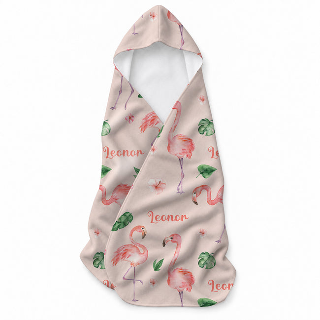 Pale blush hooded towel with pink flamingos, green tropical leaves, small flowers, and the name Leonor in coral script.