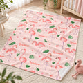 A personalized baby blanket with a pink flamingo and green leaf pattern, laid out on a cream-colored carpet in a nursery setting. The blanket features the name 'Leonor' printed in coral script alongside watercolor illustrations of pink flamingos and green leaves.