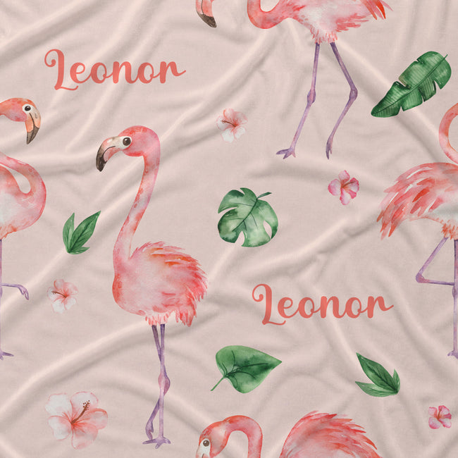 A close-up of a fabric surface featuring a repeating watercolor-style flamingo print on a light blush pink background, with green tropical leaves and coral script text reading 'Leonor'.