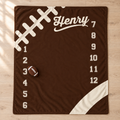 A personalized football-themed milestone blanket in dark brown with white graphics, including the name 'Henry', numbered positions 1 through 12, a football graphic, and a small brown football placed on the mat, shown on a light wood floor.