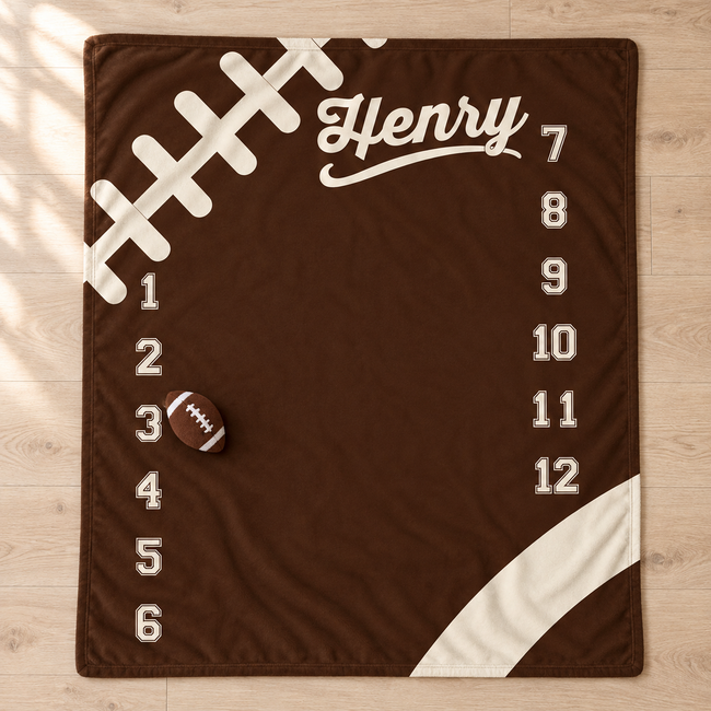 A personalized football-themed milestone blanket in dark brown with white graphics, including the name 'Henry', numbered positions 1 through 12, a football graphic, and a small brown football placed on the mat, shown on a light wood floor.