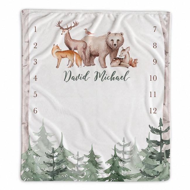 A personalized baby milestone blanket with a woodland theme, featuring watercolor illustrations of a bear, deer, fox, and pine trees on a white background. The center is personalized with the name 'David Michael' in script, and numbers 1 through 12 are printed vertically on both sides.