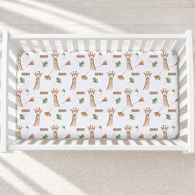 A white crib mattress fitted sheet with a repeating pattern of brown giraffes, green leaves, and the name 'ADAM' in brown text, shown inside a white wooden crib on a light textured floor.