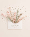A white envelope on a light cream background with scattered gold and pink confetti. A bouquet of pastel ribbons and dried floral elements bursts from the envelope, which has the words 'a gift for you' and a small heart printed in gold foil.