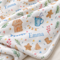 A close-up of a soft fleece blanket with a white base, featuring personalized embroidery with the name 'Emma' in blue, alongside gingerbread house, mug, stars, and berries motifs in multiple colors.