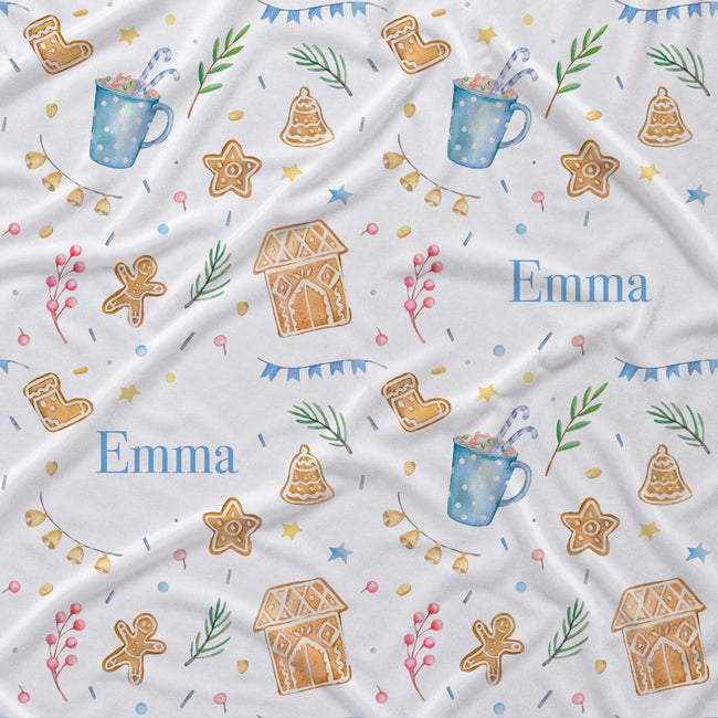 A white fabric blanket with a festive Christmas pattern featuring gingerbread cookies, hot chocolate mugs, bells, stars, and the personalized name 'Emma' in blue text.