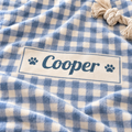 A close-up of a soft fleece blanket with a blue and white gingham check pattern, featuring a personalized cream label that reads 'Cooper' with blue paw prints on either side. A beige rope tassel is visible in the top right corner.