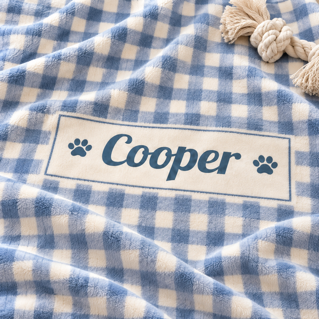 A close-up of a soft fleece blanket with a blue and white gingham check pattern, featuring a personalized cream label that reads 'Cooper' with blue paw prints on either side. A beige rope tassel is visible in the top right corner.