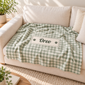 A green and white gingham dog blanket with a personalized nameplate reading 'Oreo' and paw prints, draped over a cream-colored sofa in a bright, sunlit living room.