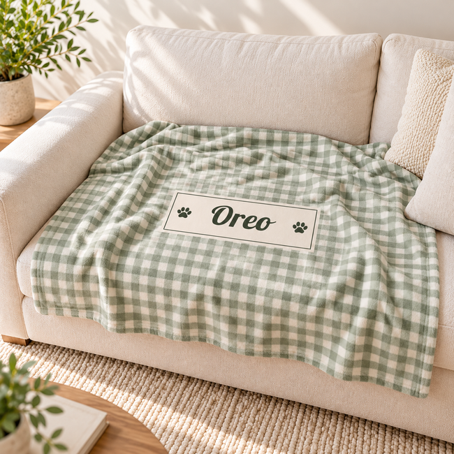 A green and white gingham dog blanket with a personalized nameplate reading 'Oreo' and paw prints, draped over a cream-colored sofa in a bright, sunlit living room.