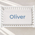 A top-down view of a white wooden crib with a fitted sheet featuring a light beige and white gingham check pattern and the name 'Oliver' printed in blue text in the center.
