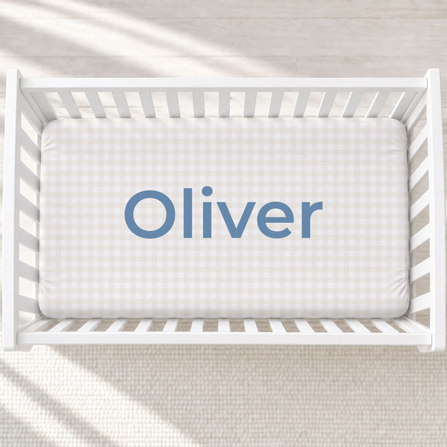 A top-down view of a white wooden crib with a fitted sheet featuring a light beige and white gingham check pattern and the name 'Oliver' printed in blue text in the center.
